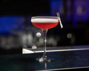 A dramatically lit, crimson cocktail with a white foam top is being lit by a bartender, creating a burst of flame from a citrus peel garnish clipped to the glass