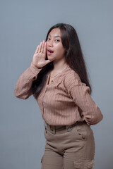 Young Indonesian woman in a beige shirt, standing with one hand near her mouth in a speaking...