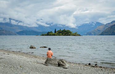 Man sitting on the rock and enjoying the view of Ruby Island. Lake Wanaka. Otago Region. South...