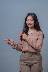 An Indonesian woman in a beige blouse pointing to her right with both hands while smiling gently....