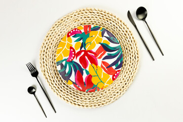 Top view of brown wicker table mat, colorful plate, black cutlery on white background. Straw round tray, exotic plant plate, summer picnic. Flat lay, copy space. 