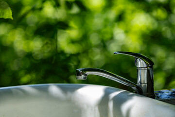 Metal faucet on a white sink outdoors with sunlight and green forest background in soft focus