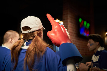 Caucasian young woman with long hair, wearing cap and oversized foam finger, cheering with group of...