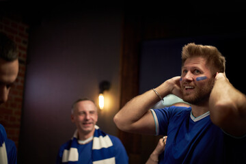 Caucasian young man with blue face paint with hands on head standing among group of soccer fans,...