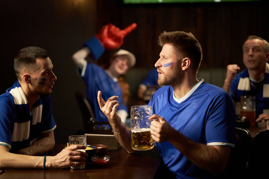 Group of excited Caucasian young men and woman wearing blue soccer jerseys cheering and talking at bar, holding drinks, celebrating soccer match, blue face paint visible on faces