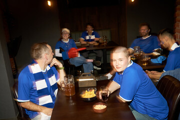 Group of young and middle aged multiethnic soccer fans sitting at table watching match, drinking beer, eating snacks, wearing blue jerseys, showing excitement and anticipation