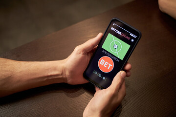 Hands of anonymous man holding smartphone displaying online soccer betting application, sitting at table in bar, engaging in sports gambling activity