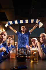 Group of Caucasian young and middle aged men watching soccer match in bar, male fan in center standing and holding scarf above head, others sitting with drinks