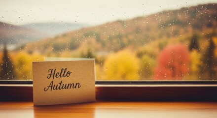 Hello autumn greeting card on a wooden sill with a rainy window and colorful fall trees outside. Autumn mood.