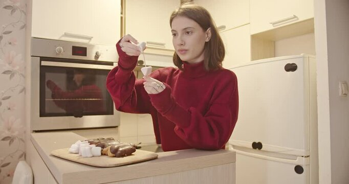 A young woman prepares a sweet treat in her kitchen. She arranges marshmallows and eclairs on skewers for a simple snack or dessert.