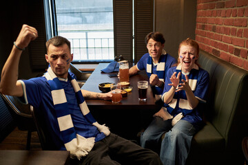 Group of young Caucasian men and woman sitting at table in sports bar cheering and celebrating football match, faces painted with team colors, drinks and snacks on table