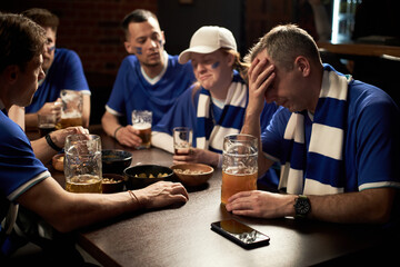 Group of Caucasian young men and woman, wearing blue team soccer jerseys, sitting at table drinking beer and reacting emotionally, while watching soccer match loss in bar