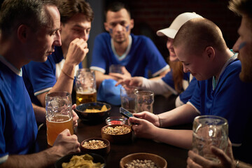 Group of happy young and middle aged Caucasian and Black men and women, wearing soccer jerseys, sitting at table watching smartphone and drinking beer, while supporting soccer team