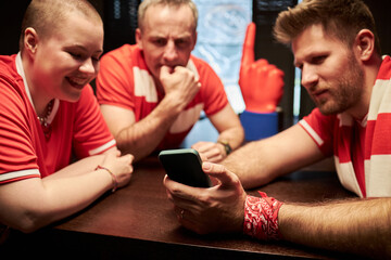 Caucasian young woman and two men watching smartphone together, wearing soccer fan attire, sitting at table in bar, engaging with mobile device during game