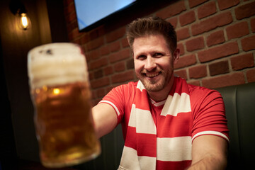 Portrait of Caucasian young man smiling and holding beer mug toward camera, wearing soccer fan scarf, sitting in sports bar with brick wall background, celebrating match