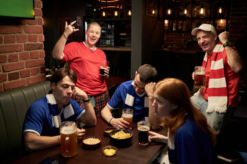 Group of multiethnic young and middle aged football fans reacting to game in bar, three Caucasian men and one woman in blue jerseys looking disappointed, two fans in red celebrating