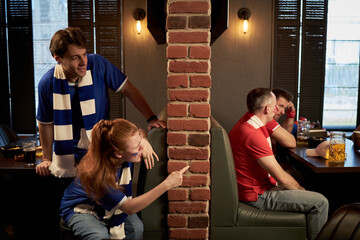 Caucasian young man and woman, wearing blue soccer scarves, leaning around brick wall, playfully interacting with two middle aged men in red jerseys, sitting at table watching football game