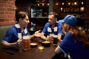 Three young soccer fans, two Caucasian men and one woman, sitting at table in sports bar talking and drinking beer, wearing matching blue jerseys and scarves, snacks on table