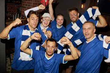 Group portrait of multiethnic sports fans celebrating, wearing blue soccer jerseys and scarves, raising fists and smiling, showing excitement while supporting their team in bar