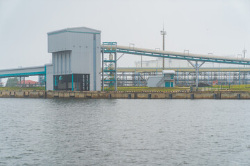 Industrial waterfront facility with conveyor system and storage building on calm riverbank,...