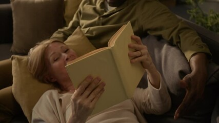 Cheerful senior Caucasian woman reading out book and laughing when lying on cushion on her African American husbands lap during cozy weekend - Powered by Adobe
