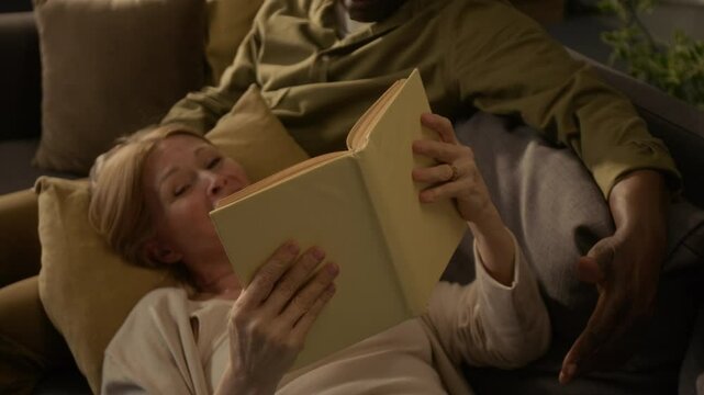 Cheerful senior Caucasian woman reading out book and laughing when lying on cushion on her African American husbands lap during cozy weekend