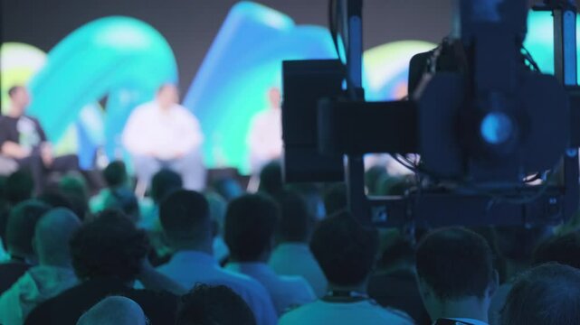 Large audience gathered for a professional presentation with a backdrop of vivid colors. The event includes speakers on stage, with a camera recording for broadcast purposes