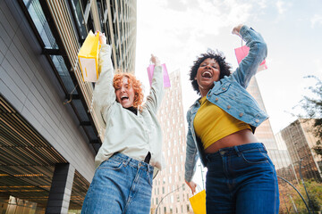 Two excited friends jubilantly celebrating their successful shopping spree with colorful bags in hand, expressing joy and happiness while raising their arms in delight during a fun day out together.