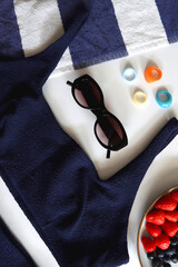 Navy swimsuit, striped beach towel, sunglasses, colorful rings and plate with berries. Top view.