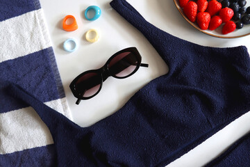 Navy swimsuit, striped beach towel, sunglasses, colorful rings and plate with berries. Top view.