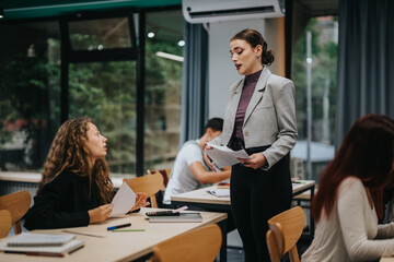 A teacher stands explaining a concept to attentive students in a modern classroom. The room is filled with natural light and learning materials. Students are seated, engaging in discussion.