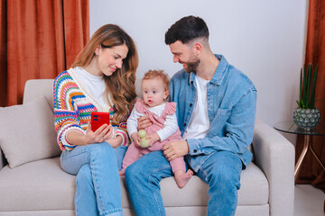 Mother shows smartphone screen to baby girl in pink overalls seated on father's lap, capturing...
