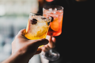 Two hands clinking cocktail glasses garnished with fruit and ice, celebrating with colorful drinks...