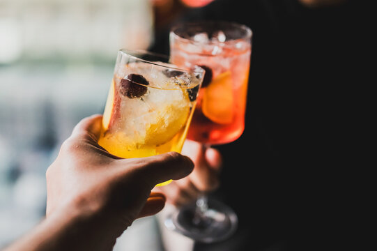 Two hands clinking cocktail glasses garnished with fruit and ice, celebrating with colorful drinks in focus and a blurred festive background. - Powered by Adobe