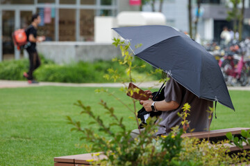 Shaded by a sun umbrella, person sits on a bench in a city park, enjoying leisure amidst natural...