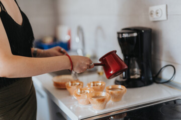 Midsection view of a woman in a modern kitchen pouring fresh coffee from a Turkish pot into small cups, with a coffee maker in the background, suggesting a morning routine.