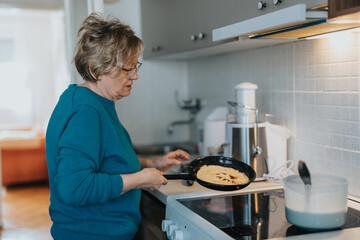 A senior woman making pancakes on a stove, surrounded by kitchen appliances, evoking feelings of...