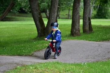Active young boy in blue jacket and helmet riding a red balance bike on an outdoor pump track....