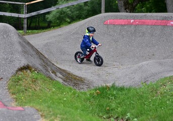 Obraz premium Active young boy in blue jacket and helmet riding a red balance bike on an outdoor pump track. Child practicing cycling skills in green park on summer day. Concept of active childhood and sport.