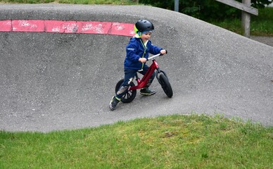 Obraz premium Active young boy in blue jacket and helmet riding a red balance bike on an outdoor pump track. Child practicing cycling skills in green park on summer day. Concept of active childhood and sport.