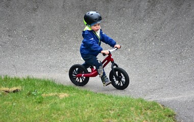 Fototapeta premium Active young boy in blue jacket and helmet riding a red balance bike on an outdoor pump track. Child practicing cycling skills in green park on summer day. Concept of active childhood and sport.