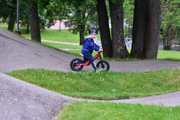 Active young boy in blue jacket and helmet riding a red balance bike on an outdoor pump track....