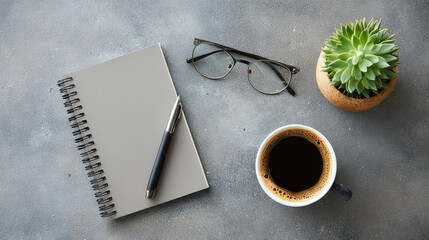 Top-down view of a workspace setup with a notebook, plant, glasses, pen, and coffee cup on a gray surface
