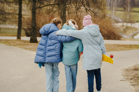 Teen girls walk together, hugging shoulders, each holding a skateboard. Jackets, jeans, sneakers show winter street style. Outdoor friendship, movement, balance, youth bonding, lifestyle and freedom