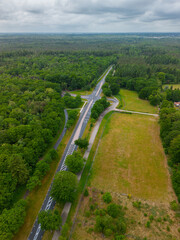 A drone view of a road cutting through dense green forest, with open grassy fields on one side and...