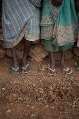 Close-up of elderly rural individuals wearing traditional clothing and sandals, standing on dry, earthy ground against a stone wall. Cultural and rustic lifestyle concept.