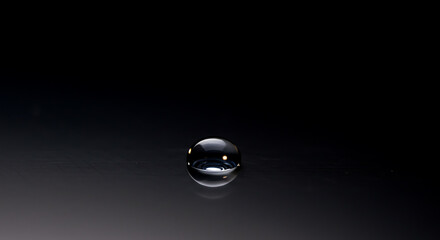A single water droplet rests on a smooth, reflective surface against a dark background.