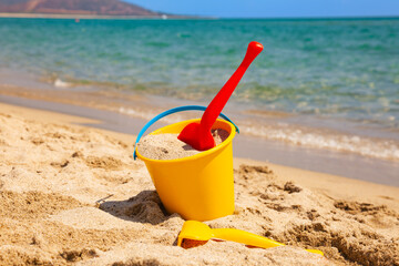 Yellow bucket with red plastic shovel filled with sand on sunny beach