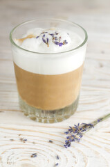 Large glass of hot Lavender Latte on a white wooden table with flowers in the background. Raf Coffee. Concept of high-quality coffee drinks. Vertical orientation. Selective focus.