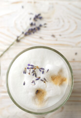 Large glass of hot Lavender Latte on a white wooden table with flowers in the background. Raf Coffee. Concept of high-quality coffee drinks. Vertical orientation. Selective focus. Top view. Copy space